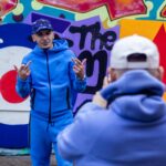 Artist Murkage posing infront of a graffiti wall in a blue tracksuit